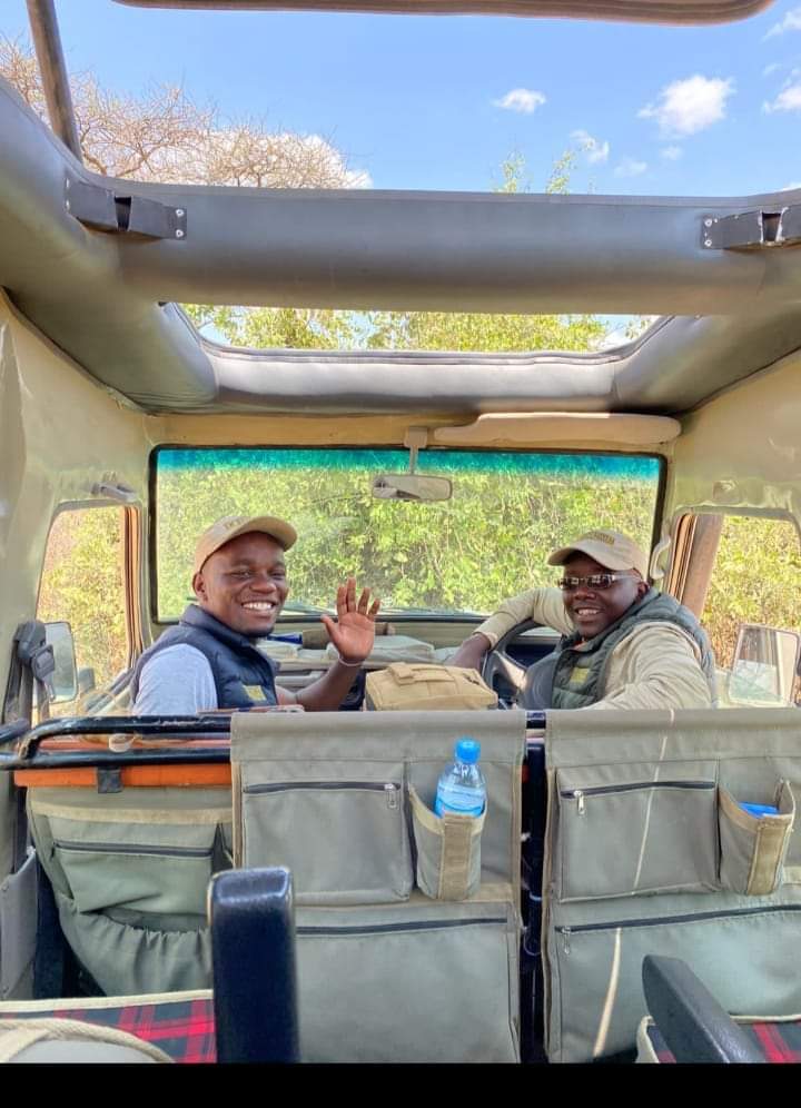 Two smiling Olympus Adventure safari guides waving from the front of a Land Cruiser